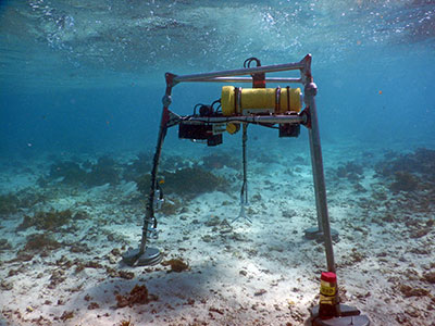 Photograph of the metal tripod frame underwater near the reef, with instruments mounted to collect data such as wave direction and currents.