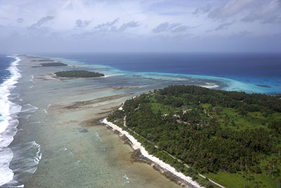 Photo taken from a plane looking along the island chain with waves breaking on beaches, trees, vegetation, and buildings on the islands, shallow waters in between the islands.