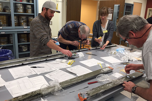 Four scientists stand around a table in a lab with gray sediment core samples on the table, and they are examining the sediment.