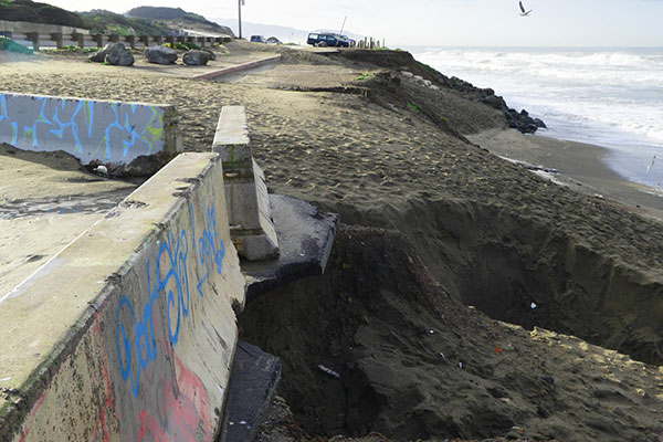 Photograph of San Francisco's Ocean Beach shows the edge of a paved parking lot with cement barracades, and the sand is eroding from under the pavement.