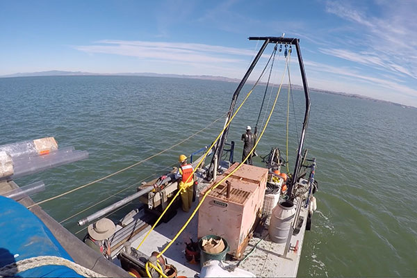 Photo taken of the stern of a boat showing scientists and equipment collecting cores of bayfloor sediment.