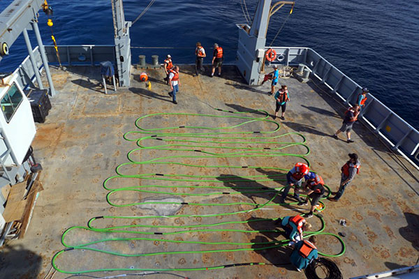 Photograph of students and a professor from the University of Washington, scientists in the Marine Geohazards group from the U.S. Geological Survey, and crew aboard the research vessel Thompson gather on the stern to haul in the seismic streamer, the long, green coil on the deck.